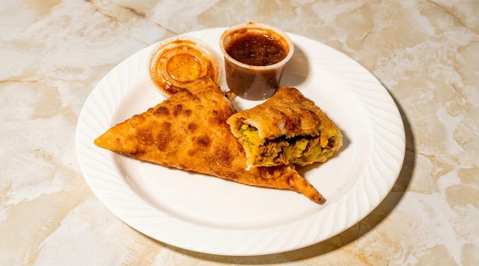 Two golden-brown samosas, one cut open to reveal a savory filling, served with two dipping sauces.