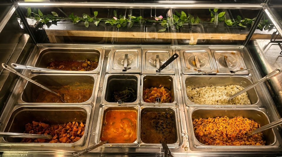 A buffet line with various dishes in metal trays, including rice, beans, and stews.